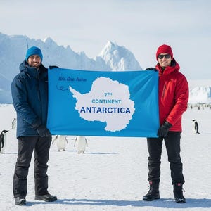 May include: Two people holding a blue flag with the text "7th Continent Antarctica" and a map of Antarctica. The background features snow, penguins, and icebergs. The people are wearing winter clothing.