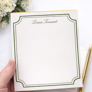 Può includere: Carta da lettere personalizzata con bordo verde e il nome "Louise Tennant" in corsivo. Una penna dorata è appoggiata accanto alla carta. Un mazzo di fiori bianchi è visibile sullo sfondo.
