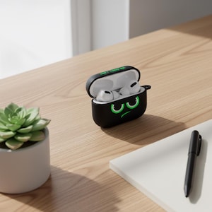 Puede incluir: Funda negra para AirPods Pro con un diseño de cara gruñona verde de dibujos animados. La funda está abierta, revelando los AirPods blancos. La parte superior de la funda tiene el texto "Don't Touch Me". También se ve una planta suculenta en una maceta blanca y un bolígrafo negro.
