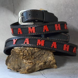 Puede incluir: Cinturón de cuero negro con la palabra "YAMAHA" en letras rojas. El cinturón tiene una hebilla plateada y se muestra sobre un trozo de madera. Accesorio de motocicleta.