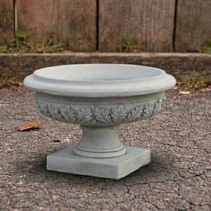 May include: A light gray concrete pedestal planter with a wide, circular basin and a square base. The exterior of the bowl features a decorative relief pattern of repeating oak leaves encircling the entire circumference of the vessel.