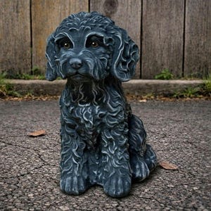 May include: A dark gray dog statue with a curly coat, sitting upright. The statue has detailed fur texture and brown eyes. The dog is positioned on a paved surface, with a wooden fence in the background.