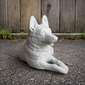 May include: A light gray, stone-like sculpture of a German Shepherd dog in a relaxed, lying down pose. The dog sculpture has detailed features, including pointed ears and a textured surface. The sculpture is placed on a cracked asphalt surface.