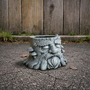 May include: A gray, tree stump-shaped planter with a sculpted face, closed eyes, and a long beard. The planter has a textured surface with mushroom and leaf details. It sits on a paved surface with a wooden fence in the background.