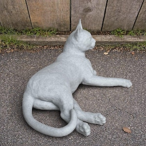 May include: A light gray cat statue in a relaxed pose, lying on a paved surface. The cat's tail curves around its legs, and one paw is extended. The statue is positioned in front of a wooden fence.