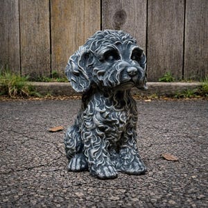 May include: A gray, textured dog statue in a seated position. The dog has curly fur and floppy ears. The statue is outdoors on a cracked asphalt surface, with a wooden fence in the background.