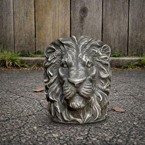 May include: A gray, detailed sculpture of a lion's head. The lion's mane is textured and the face has intricate features. The sculpture is placed on a dark, cracked surface, with a wooden fence in the background.