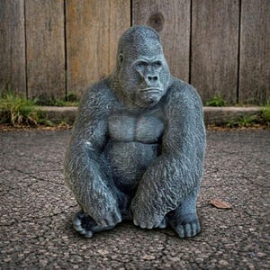 May include: A gray gorilla statue in a seated position, with a detailed texture. The gorilla has a muscular build and is positioned on a cracked asphalt surface. The background features a wooden fence and some grass.