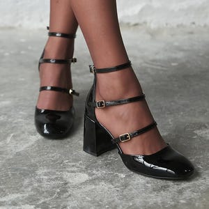 May include: Black patent leather block heel shoes featuring a square toe and three thin adjustable ankle straps with small gold-toned metal buckles. The shoes have a glossy finish and a sturdy, thick heel, providing a structured look for the feet.