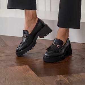 May include: Black leather loafers with a chunky lug sole and a gold-tone horsebit detail. The shoes have a fringed vamp and a block heel. The loafers are worn with black pants on a wood floor.