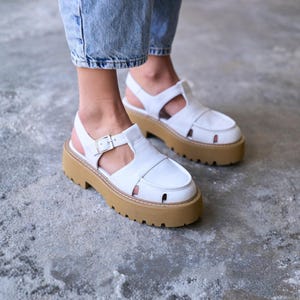 May include: White leather fisherman sandals featuring a closed-toe design with side cutouts, a buckled ankle strap, and a thick, tan-colored lug sole. The footwear has a chunky platform base with deep treads and visible stitching along the midsole.