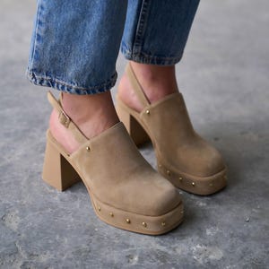 May include: Tan suede clogs with a square toe and a chunky heel. The open-back shoes have a strap with a buckle and are adorned with small gold-colored studs around the sole.