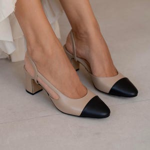 Puede incluir: Zapatos de tacón slingback beige y negro con tacón de bloque. Los zapatos tienen punta afilada y una correa alrededor del tobillo. Los tacones miden aproximadamente 6,35 cm de alto.