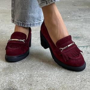 May include: Burgundy suede loafers with a silver-tone buckle and fringe detail. The shoes have a chunky black heel and sole. The loafers are worn with light wash denim jeans.