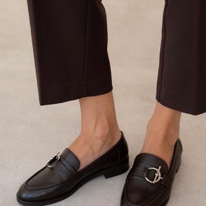 May include: A pair of black leather loafers with silver-tone hardware. The shoes have a classic design with a rounded toe and a strap across the top. The image shows the shoes being worn with dark brown pants.