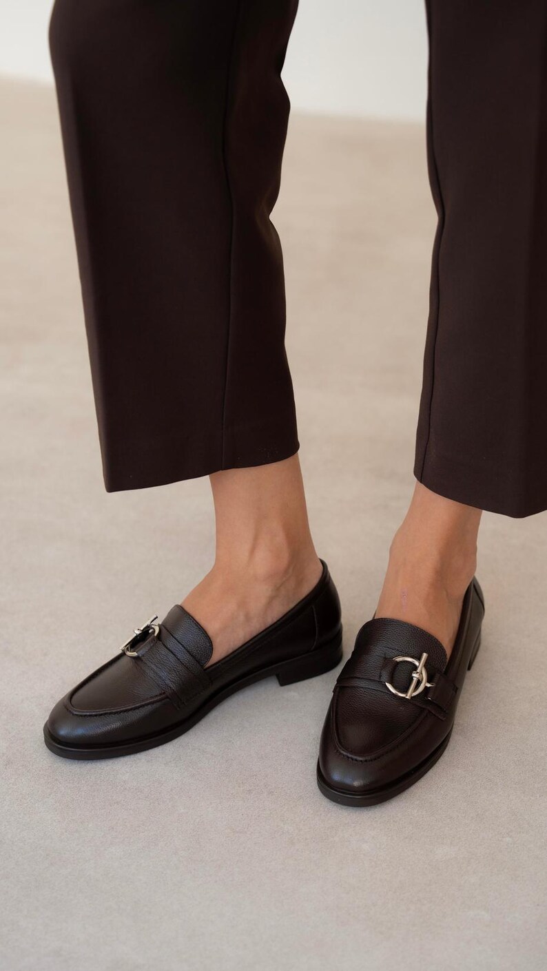 May include: A pair of black leather loafers with silver-tone hardware. The shoes have a classic design with a rounded toe and a strap across the top. The image shows the shoes being worn with dark brown pants.