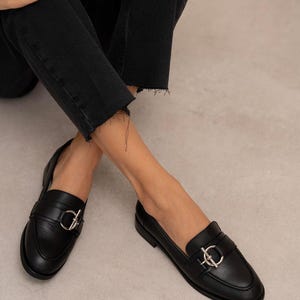 May include: Black leather loafers with silver-colored hardware. The shoes have a classic design with a rounded toe and a decorative buckle. The image shows the shoes from a low angle.