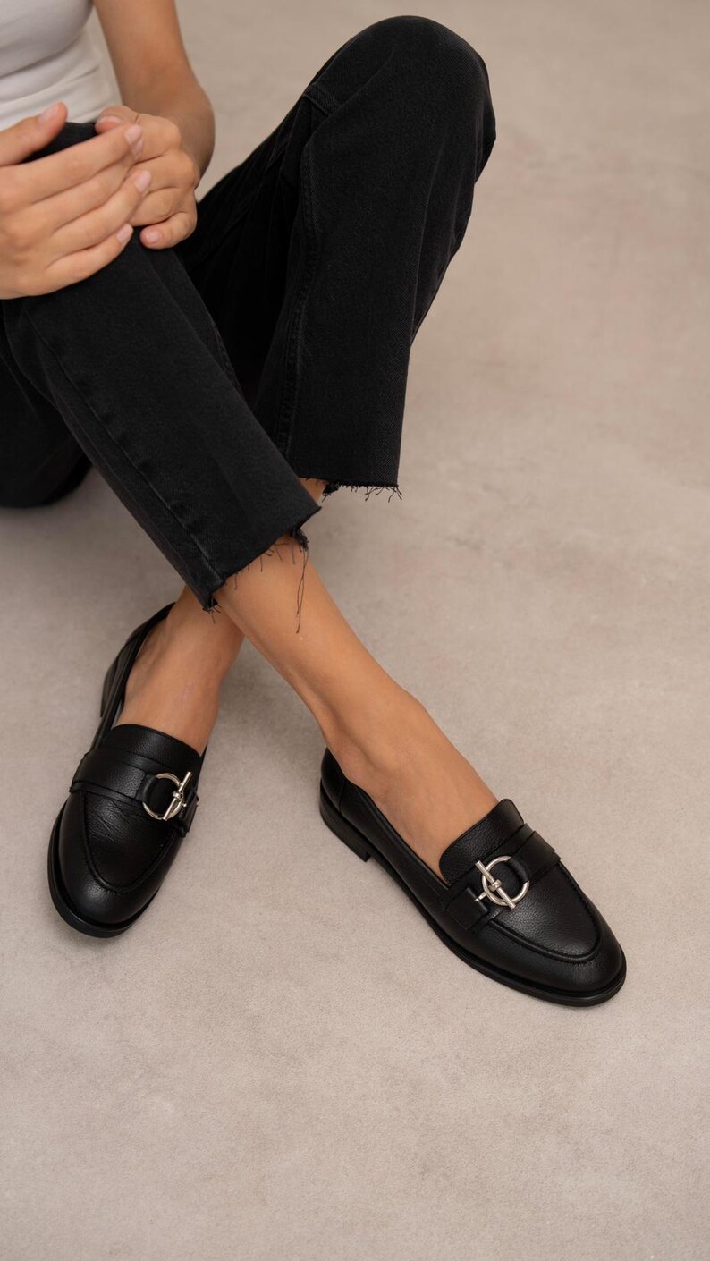 May include: Black leather loafers with silver-colored hardware. The shoes have a classic design with a rounded toe and a decorative buckle. The image shows the shoes from a low angle.