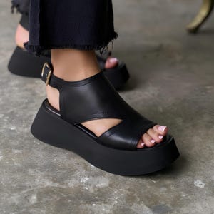 May include: Black platform sandals with a peep-toe design and a gold-colored buckle. The sandals have a thick sole and a cutout detail on the upper. The footwear is made of a smooth material.