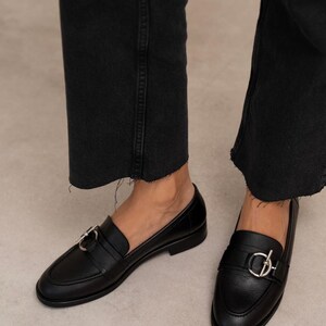 May include: Black leather loafers with silver-tone hardware. The shoes have a classic design with a rounded toe and a strap across the top. The loafers are worn with black denim jeans.