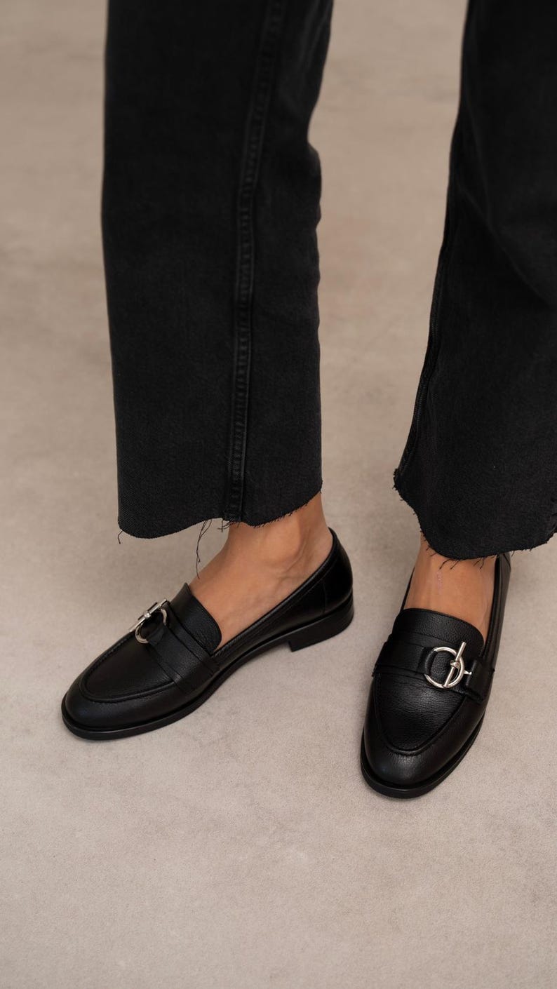 May include: Black leather loafers with silver-tone hardware. The shoes have a classic design with a rounded toe and a strap across the top. The loafers are worn with black denim jeans.