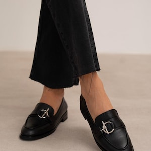 May include: Black leather loafers with a silver-colored buckle detail. The shoes have a rounded toe and a low heel. The image shows the shoes from a low angle, with the wearer's legs visible in black denim jeans.