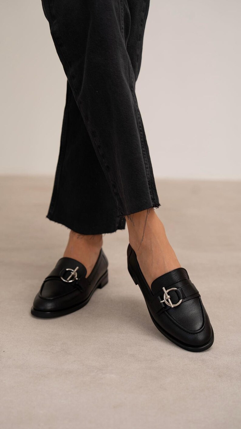 May include: Black leather loafers with a silver-colored buckle detail. The shoes have a rounded toe and a low heel. The image shows the shoes from a low angle, with the wearer's legs visible in black denim jeans.