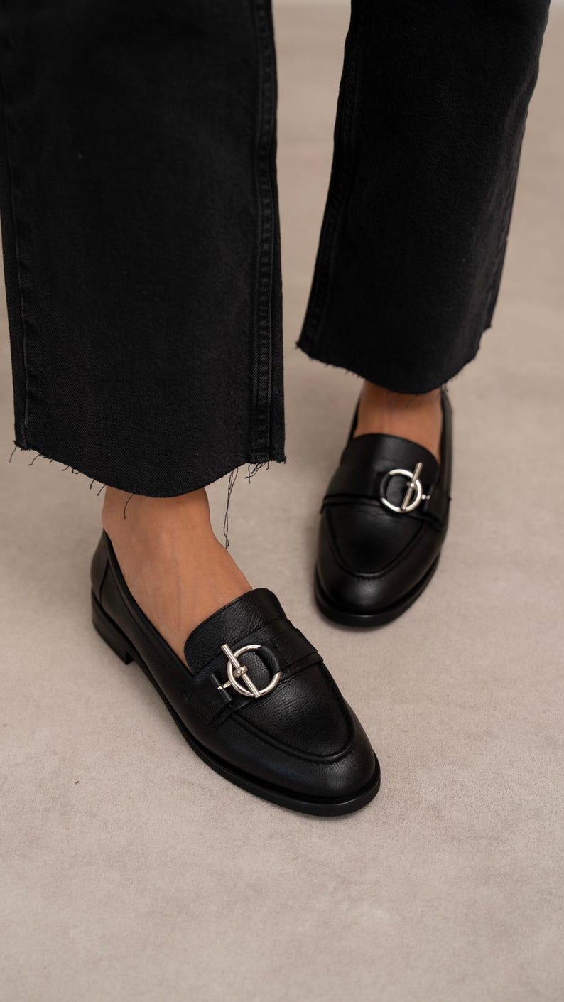 May include: Black leather loafers with silver-tone hardware. The shoes have a classic slip-on design with a rounded toe and a decorative buckle detail. The loafers are paired with black denim jeans.