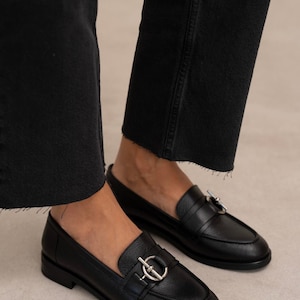 May include: A pair of black leather loafers with a silver-colored metal accent on the vamp. The shoes have a rounded toe and a low heel. The image shows the shoes from a low angle, with the wearer's black jeans visible.