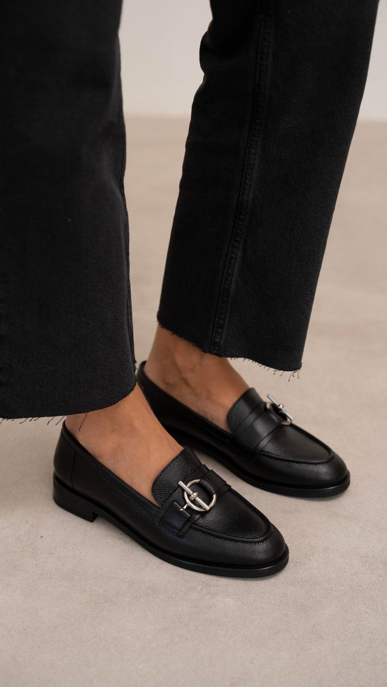 May include: A pair of black leather loafers with a silver-colored metal accent on the vamp. The shoes have a rounded toe and a low heel. The image shows the shoes from a low angle, with the wearer's black jeans visible.