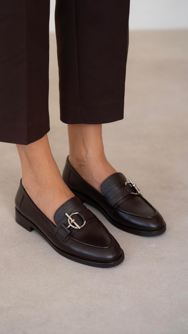 May include: A pair of dark brown leather loafers with silver-colored hardware. The shoes have a classic design with a rounded toe and a low heel. The loafers are worn with dark brown trousers.
