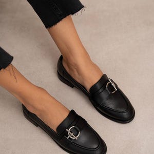 May include: Black leather loafers with a silver-colored buckle detail. The shoes have a classic design with a rounded toe and a low heel. The image shows the shoes being worn, with the focus on the footwear.