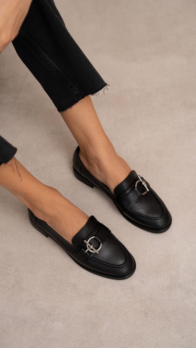 May include: Black leather loafers with a silver-colored buckle detail. The shoes have a classic design with a rounded toe and a low heel. The image shows the shoes being worn, with the focus on the footwear.