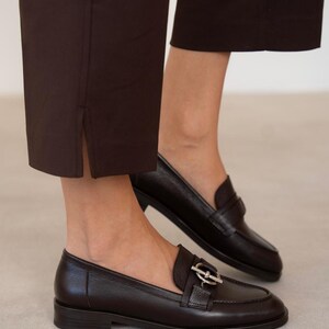 May include: Dark brown leather loafers with a silver buckle detail. The shoes are paired with dark brown trousers. The loafers have a classic design with a low heel and a rounded toe. The trousers have a straight leg and a slit at the ankle.