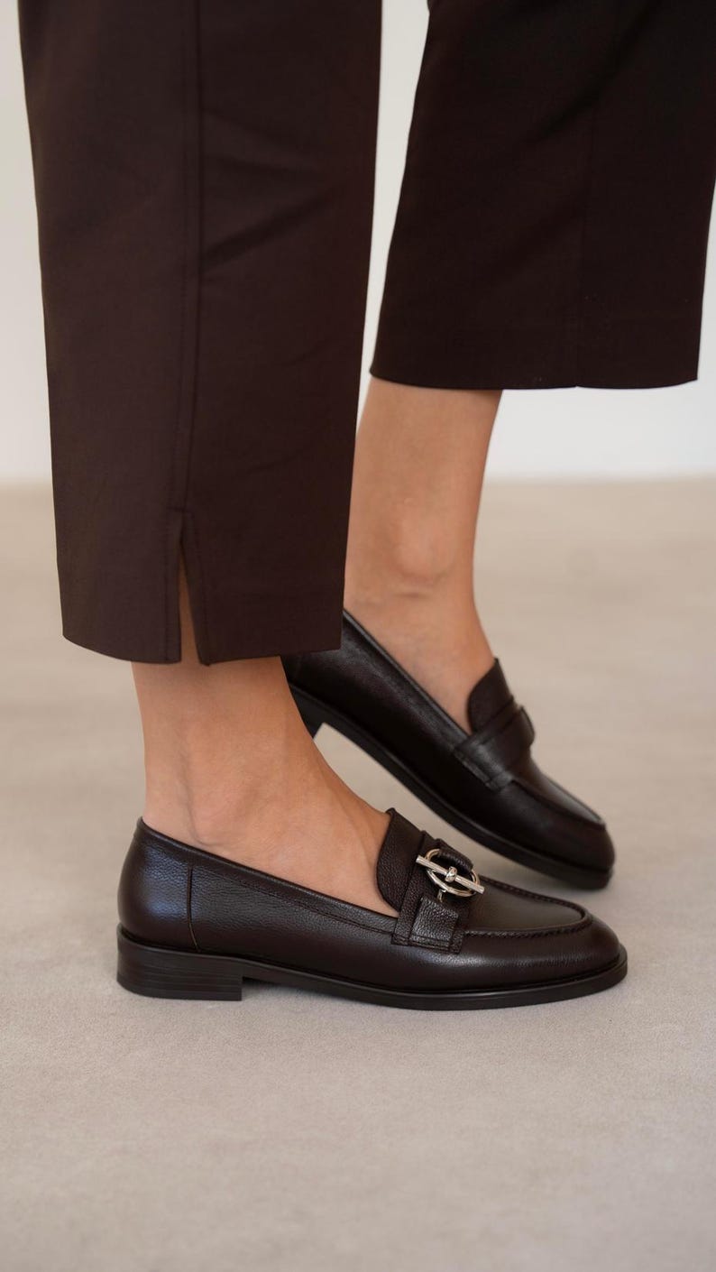 May include: Dark brown leather loafers with a silver buckle detail. The shoes are paired with dark brown trousers. The loafers have a classic design with a low heel and a rounded toe. The trousers have a straight leg and a slit at the ankle.
