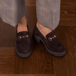 May include: Dark brown suede loafers with a gold-colored horsebit detail and fringe. The shoes have a chunky, dark gray sole. The image shows the shoes on a wooden floor, with gray pants visible.