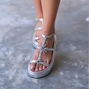May include: A pair of silver platform sandals featuring a T-strap design with two decorative star-shaped embellishments adorned with clear crystals. The sandals have a metallic finish, an adjustable ankle strap with a buckle, and a thick platform sole.