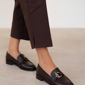 May include: A pair of dark brown leather loafers with a silver-colored buckle detail. The shoes are paired with dark brown cropped trousers. The image is taken from a low angle, showing the shoes and the lower legs.