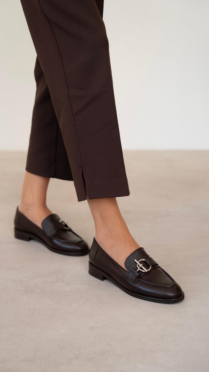 May include: A pair of dark brown leather loafers with a silver-colored buckle detail. The shoes are paired with dark brown cropped trousers. The image is taken from a low angle, showing the shoes and the lower legs.