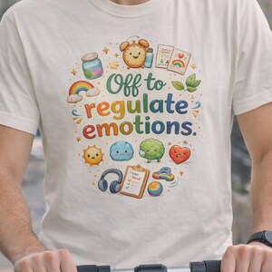 May include: A cream-coloured t-shirt featuring the text "Off to regulate emotions" in a colourful design. The graphic includes a rainbow, sun, cloud, broccoli, and heart. The t-shirt is being worn.