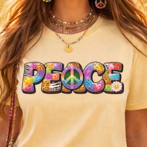 Könnte beinhalten: Ein hellgelbes T-Shirt mit dem Wort "PEACE" in bunten, gemusterten Buchstaben. Die Buchstaben weisen eine Vielzahl von Designs auf, darunter Batik, Tiermuster und ein Friedenszeichen. Eine Gänseblümchen und ein Smiley sind ebenfalls enthalten.