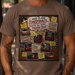 以下が含まれることがあります： 陰謀論に関するグラフィックデザインが施されたトープ色のTシャツ。「I NEED A NEW CONSPIRACY... THEY'RE ALL BEING PROVEN TRUE!」のテキストと画像、そして「CONFIRMED」の文字が含まれています。