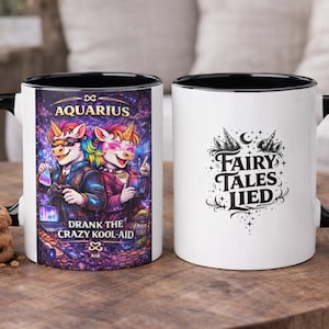 May include: Two white ceramic mugs with black handles and rims. One mug features a colourful illustration of unicorn-like creatures with the text "Aquarius" and "Drank the Crazy Kool-Aid." The other mug has the text "Fairy Tales Lied."