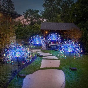 May include: Four solar-powered garden lights with a firework design. Each light features a cluster of colorful LED lights radiating outwards from a central point. The lights are installed along a stone pathway in a garden setting.