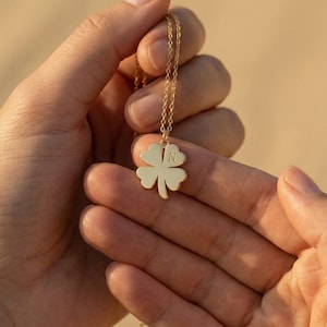 May include: A gold-colored necklace featuring a four-leaf clover pendant. The pendant has the letter "N" engraved on it. The necklace is held in a person's hands against a blurred background.