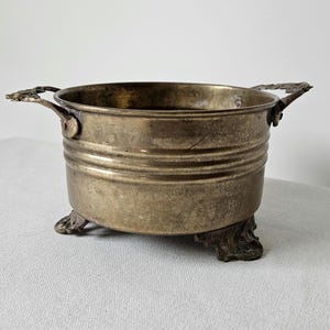 May include: A vintage brass planter with two ornate handles and three decorative feet. The planter has a round shape with horizontal ridges around the body. The brass has a tarnished, aged appearance, suggesting it is an antique or vintage piece.