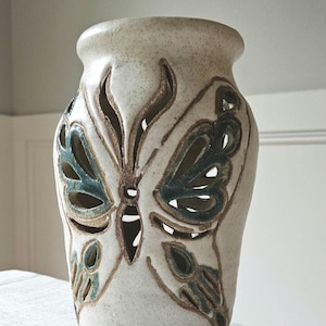May include: A cream-colored ceramic vase with a butterfly design. The butterfly is cut out of the vase, with teal and brown accents. The vase is approximately 10 inches tall and has a rounded shape.