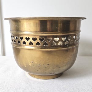 May include: A vintage brass planter with a heart-shaped cut-out design around the middle. The planter has a rounded base and a slightly flared rim. The brass has a warm, aged patina, suggesting it is an antique.