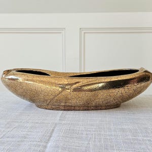 May include: A gold-colored, oval-shaped ceramic vase with a cracked, textured surface. The vase has two openings and a dark interior. The decorative piece sits on a light-colored surface.