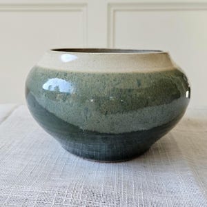 May include: A handcrafted ceramic bowl with a wide, rounded shape. The bowl features a gradient of colors, transitioning from a light beige rim to shades of green and teal. The glossy finish reflects light, highlighting the smooth surface.